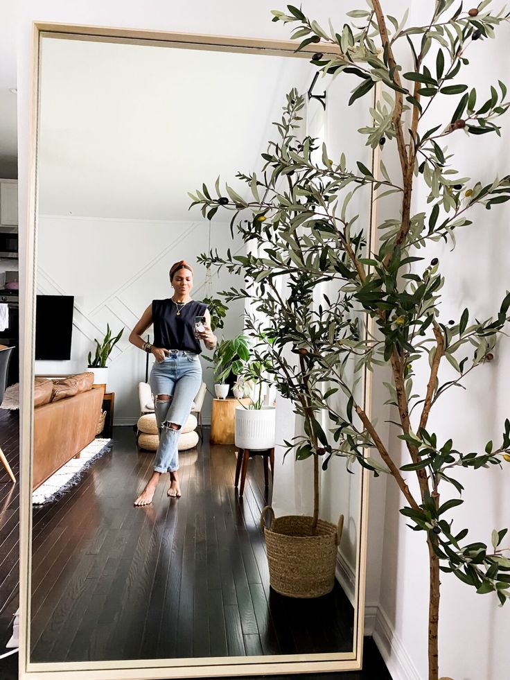 a woman standing in front of a mirror next to a potted tree and couch