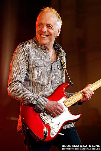 a man playing a red electric guitar on stage at a music festival, smiling and holding it in his right hand