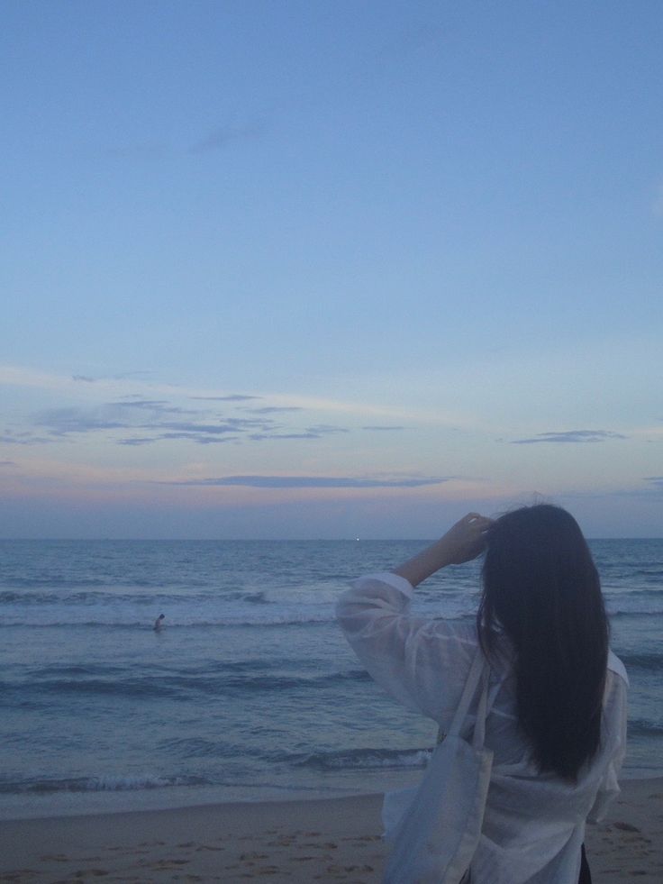 a woman standing on top of a beach next to the ocean holding her hair back