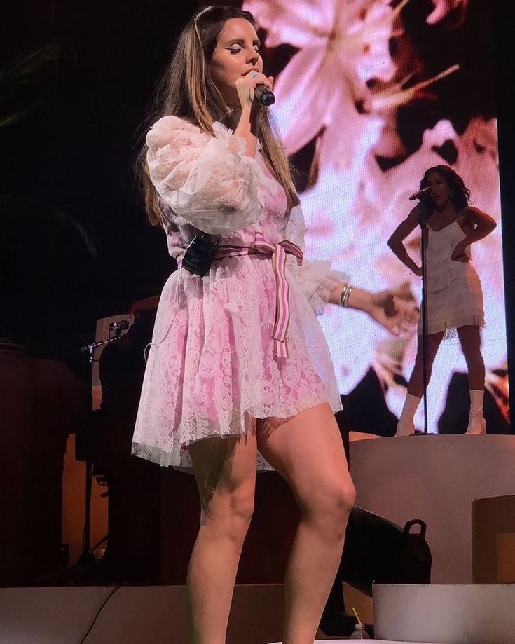 a woman standing on top of a stage with a microphone in her hand and wearing a pink dress