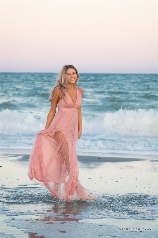 a woman in a pink dress is standing on the beach at sunset with her hair blowing in the wind