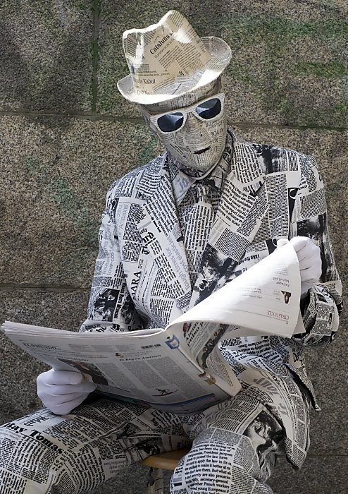 a man in a newspaper suit and hat sitting on a bench reading a paper while wearing sunglasses