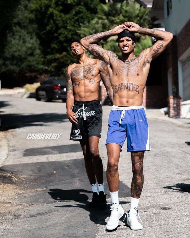 two men walking down the street with tattoos on their body and arms, one man has his hands behind his head