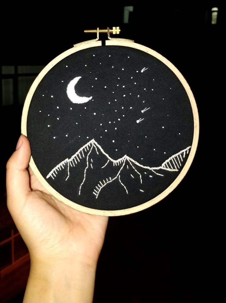 a hand holding up a black and white embroidery hoop with mountains, stars and the moon