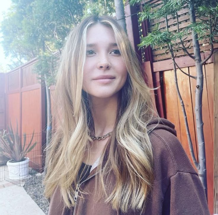 a woman with long hair standing in front of a wooden fence and wearing a brown hoodie