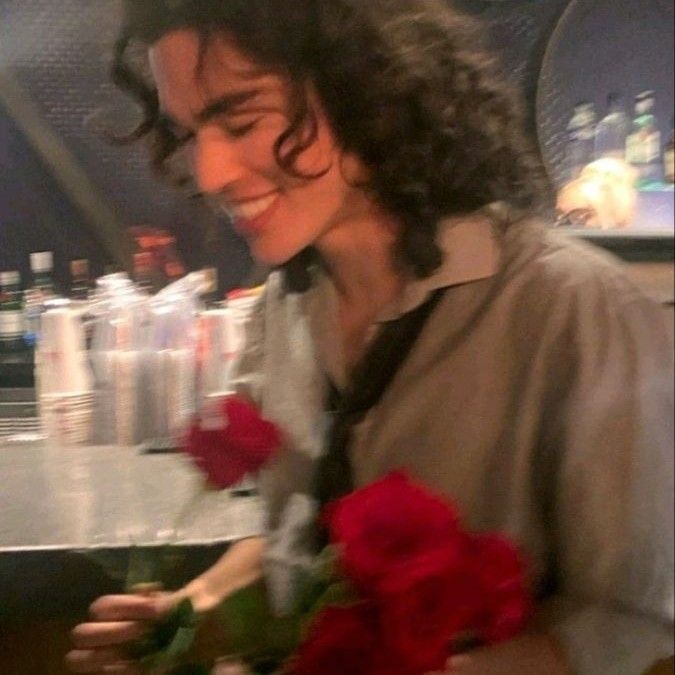 a man with long curly hair holding flowers in his hand and smiling at the camera