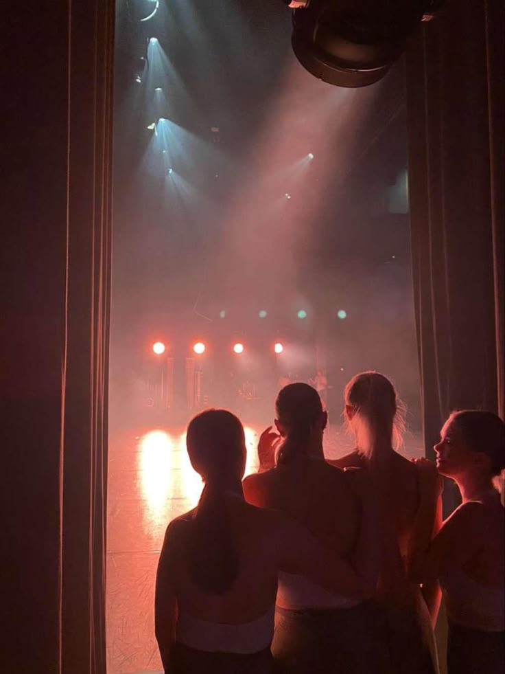 three women standing in front of a stage with lights on it and their arms around each other