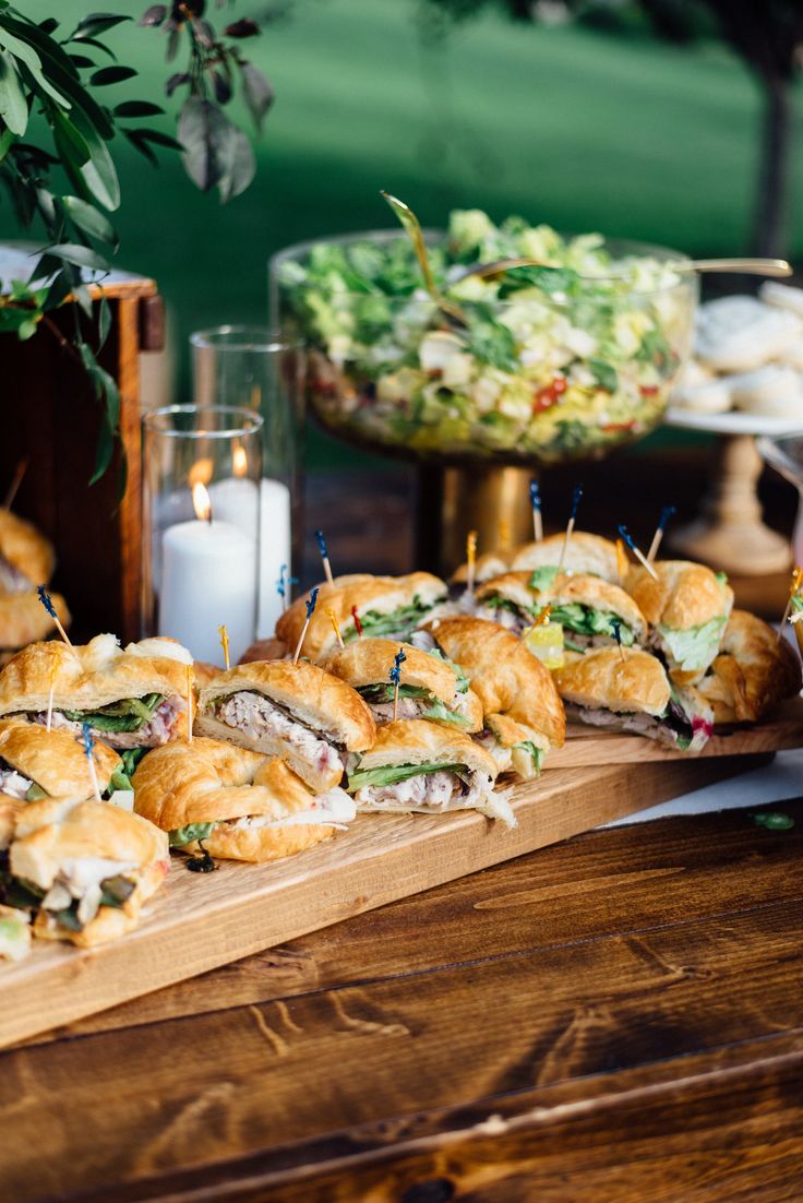 many sandwiches are arranged on a wooden board with toothpicks in the middle and some other food behind them