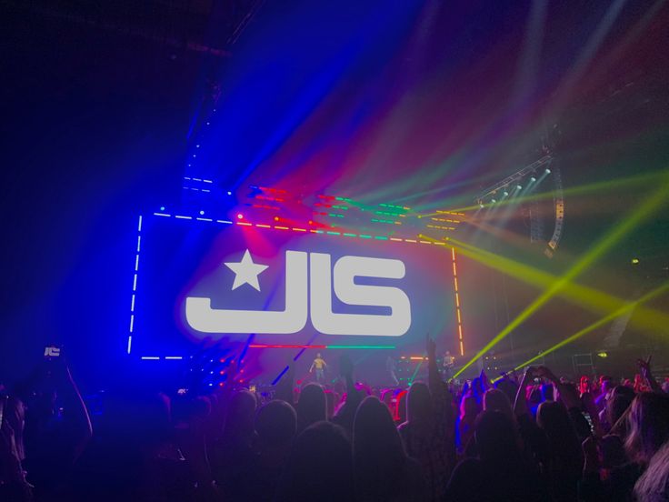 a large group of people standing on top of a stage under bright colored lights at a concert