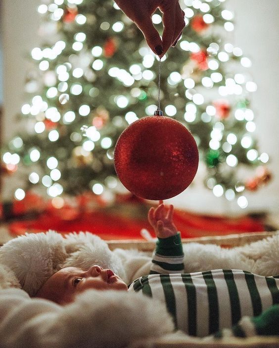 a baby is laying in front of a christmas tree with a red ornament hanging from it's head