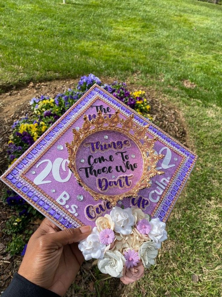 someone holding up a purple and gold graduation cap with flowers on it in front of some grass