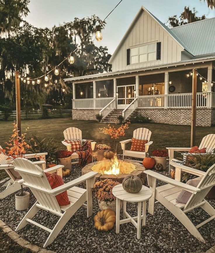 a fire pit surrounded by chairs and pumpkins in front of a white house with lights strung from the porch