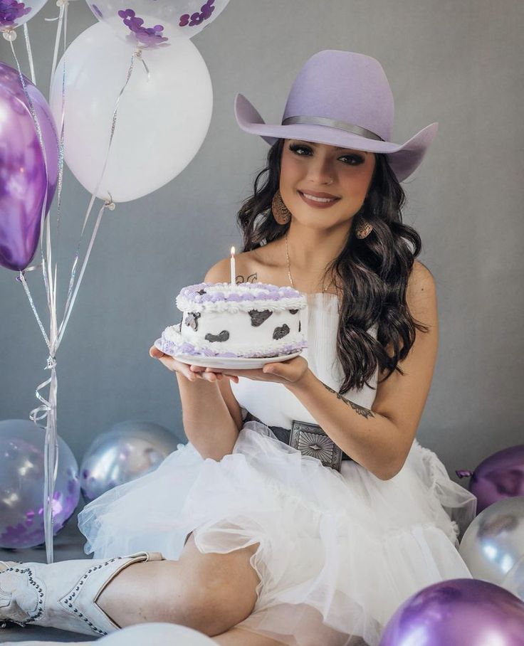 a woman in a dress and hat holding a cake with candles on it while sitting next to balloons