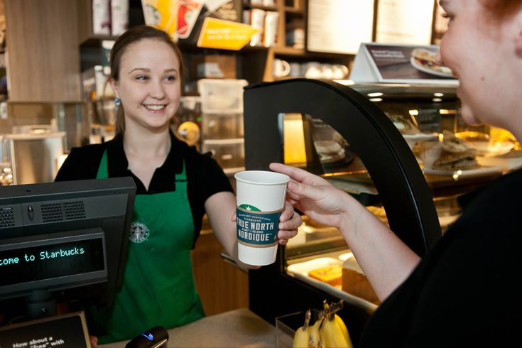 a woman holding up a cup of coffee in front of a man behind the counter