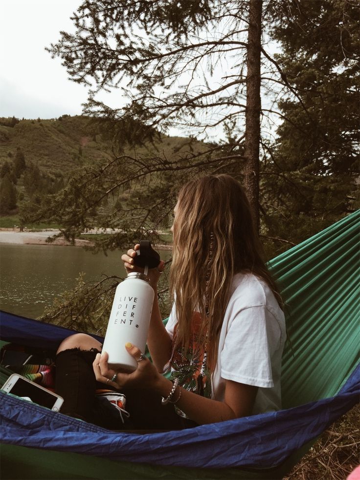 a woman sitting in a hammock holding a bottle and looking at the water