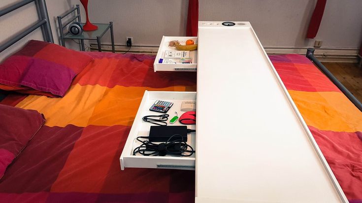 a bed with a red and orange bedspread next to an electronic device on top of it