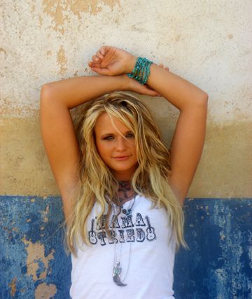 a beautiful young blond woman leaning against a wall with her hands on her head and looking at the camera