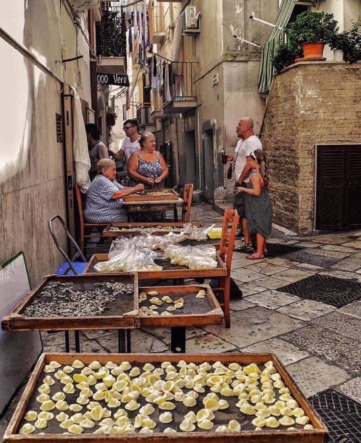 several people sitting at tables in an alleyway with food on the table and one person standing next to them