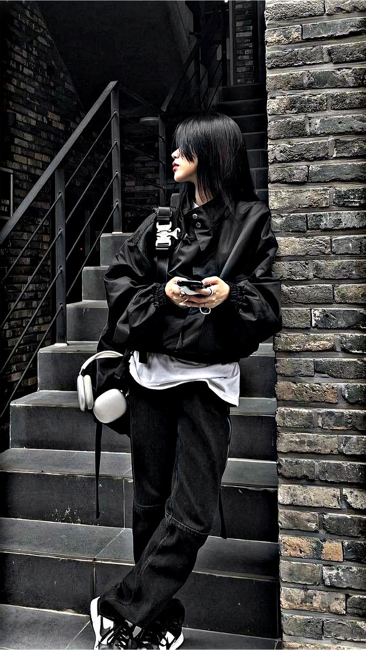 a woman with black hair standing on the stairs looking at her cell phone while wearing all black