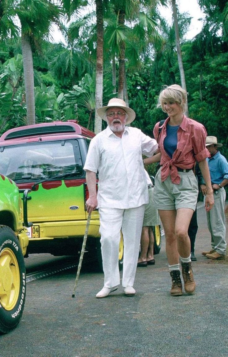 an older man and woman walking in front of a green truck with yellow wheels on the road