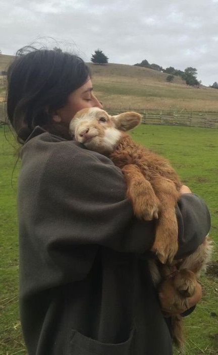 a woman holding a baby goat in her arms while standing on top of a lush green field