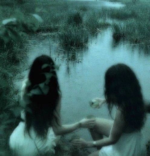 two young women sitting on the ground near a body of water, one with her hands in her other's lap