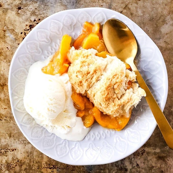 a white plate topped with ice cream and peach cobbler next to a gold spoon