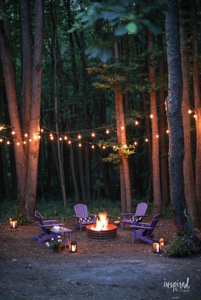 a fire pit surrounded by chairs in the woods with lights strung over it at night