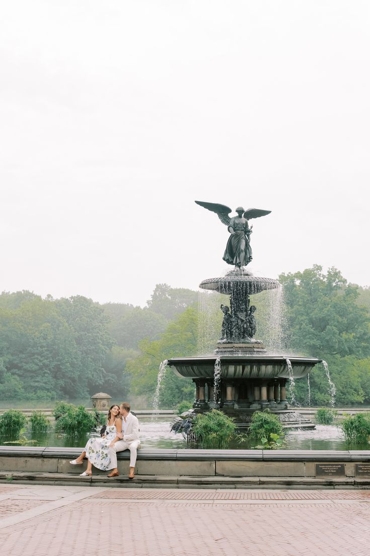 two people sitting on a bench in front of a fountain with an angel statue behind them