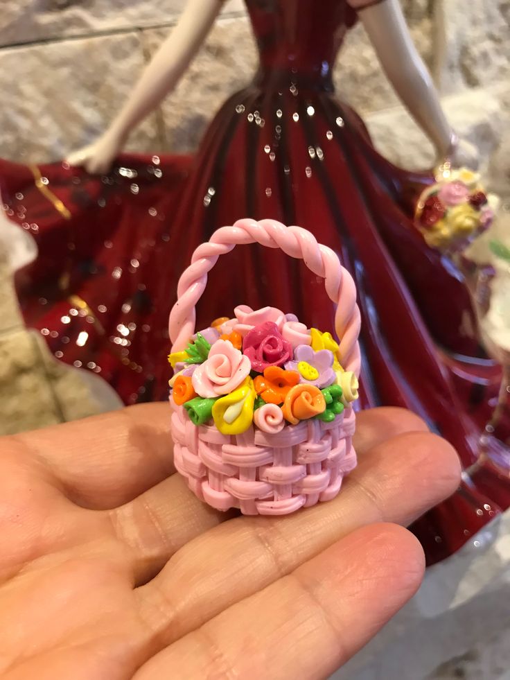 a hand holding a small pink basket with flowers on it and a doll in the background