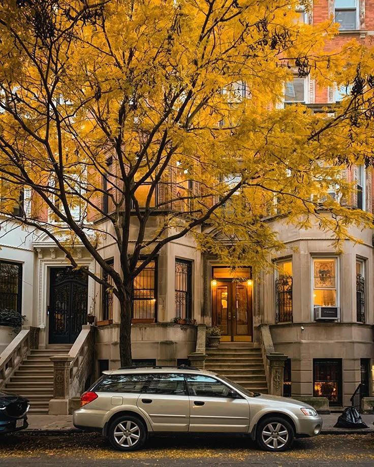 two cars parked in front of a building with yellow leaves on the tree and stairs leading up to it