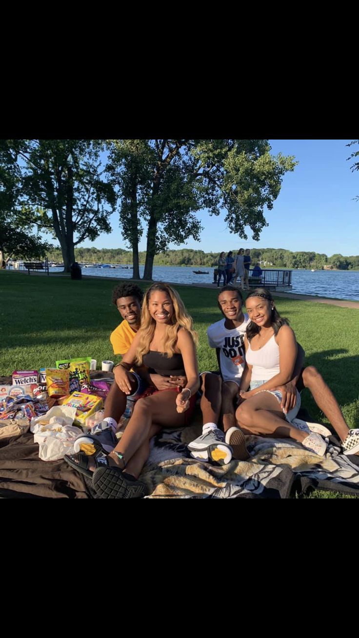 four people sitting on a blanket in the grass with food and drinks laid out around them