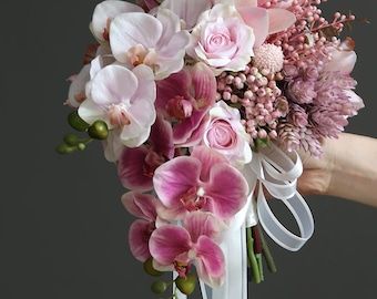 a bridal bouquet with pink flowers and white ribbon tied around the center, held by a woman's hand