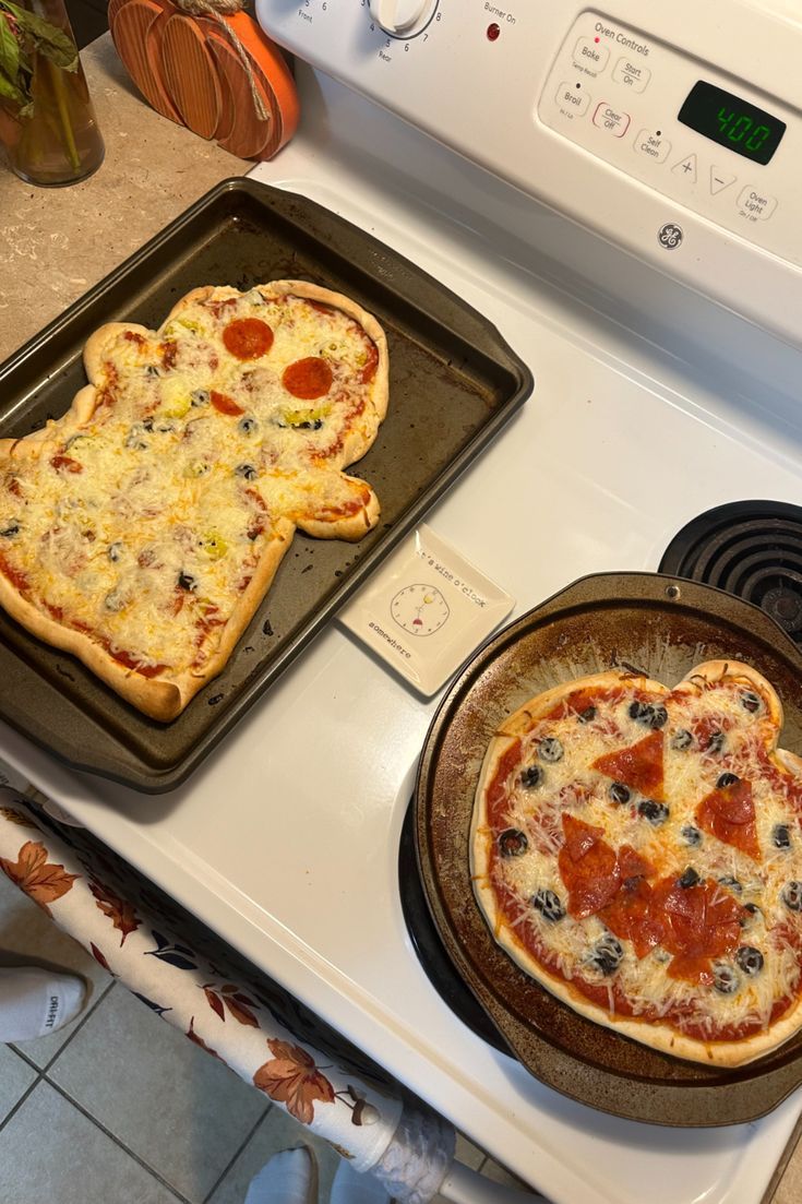 two pizzas on pans sitting on top of an oven with one being shaped like a heart