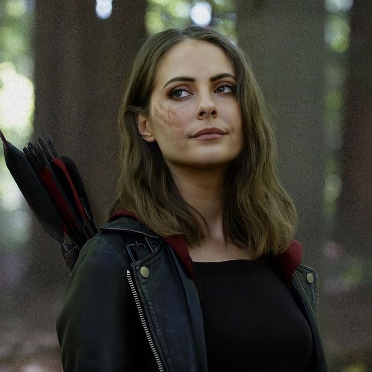 a woman holding an arrow in her hand and looking up at the sky with trees behind her