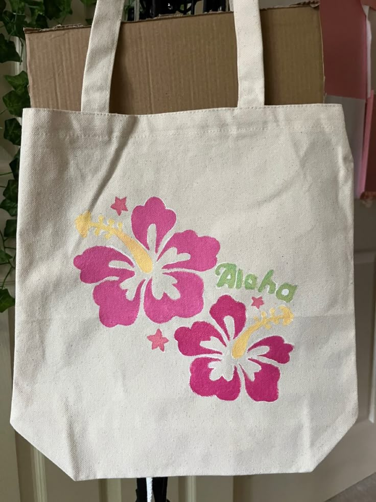 a white tote bag with pink and yellow flowers painted on it, hanging from a hook