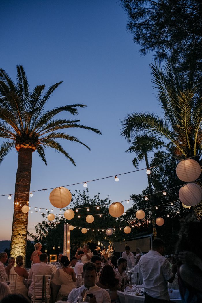 a group of people sitting at tables under paper lanterns with palm trees in the background