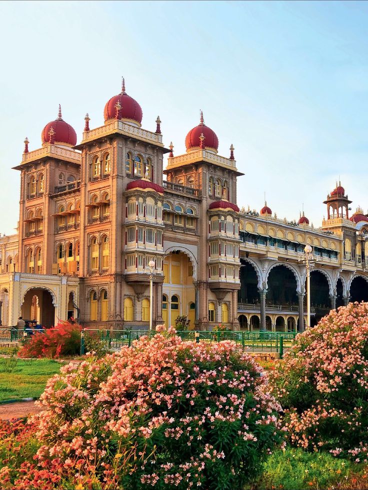 an ornate building with red domes on top and pink flowers in the foreground, surrounded by greenery