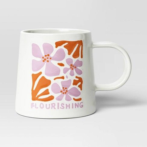 a white coffee mug with an orange and pink flower design on the front, which says flourishing