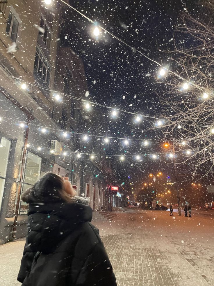 a person standing on a snowy street looking up at the sky with lights strung overhead