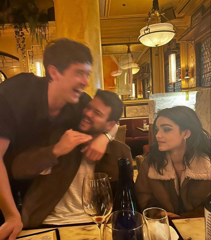 two people sitting at a table with wine glasses in front of them and one person standing up