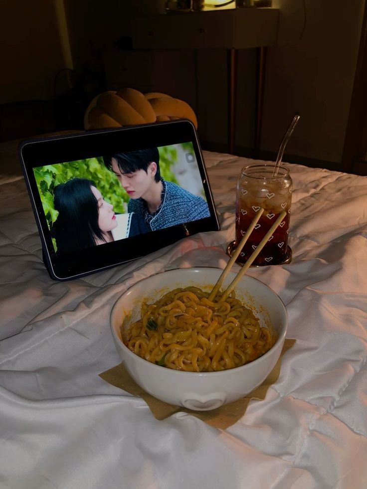 a bowl of noodles with chopsticks in front of an ipad on a bed