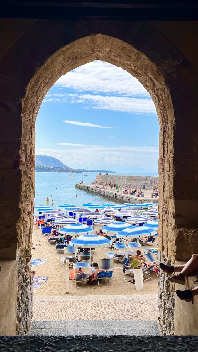 an archway leading to the beach with blue umbrellas and people on the water in the distance