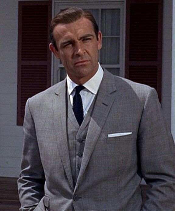 a man wearing a suit and tie standing in front of a house with shutters