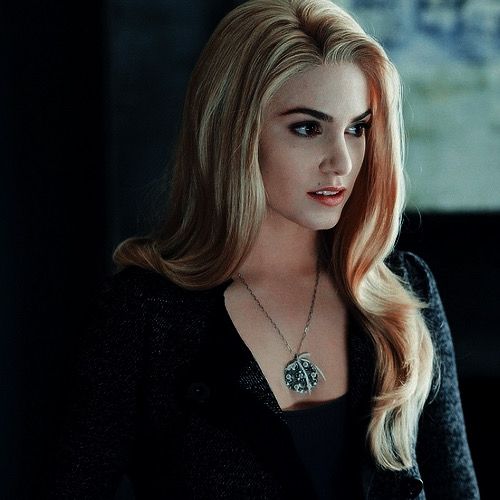 a woman with long blonde hair wearing a black jacket and necklace in a dark room