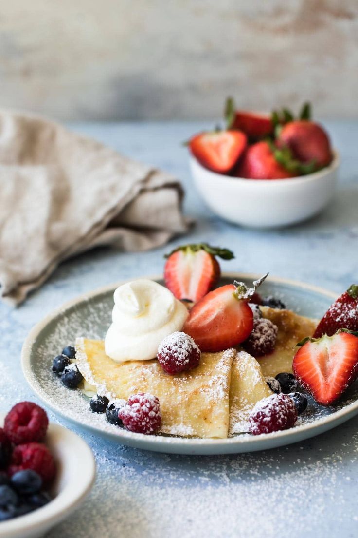 a white plate topped with pancakes covered in powdered sugar and strawberries next to bowls of berries