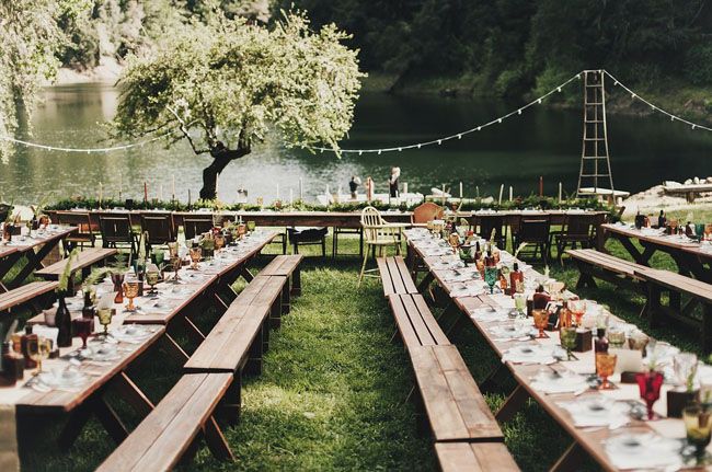 a long table set up for an outdoor dinner by the river with lights strung over it