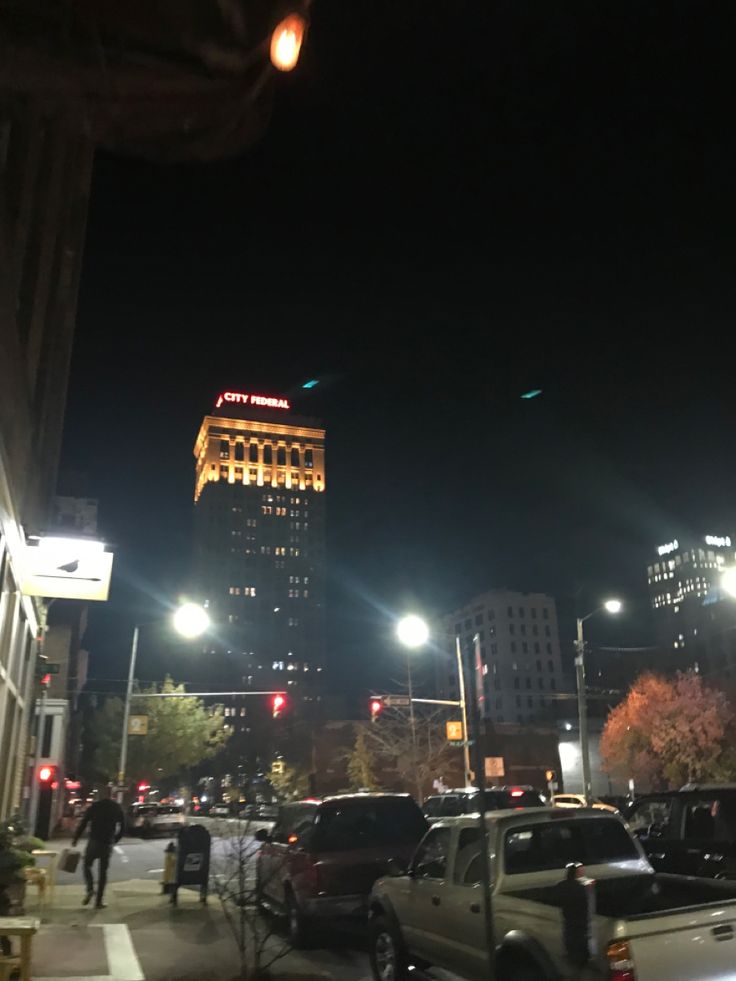 cars are parked on the side of the road at night in front of tall buildings