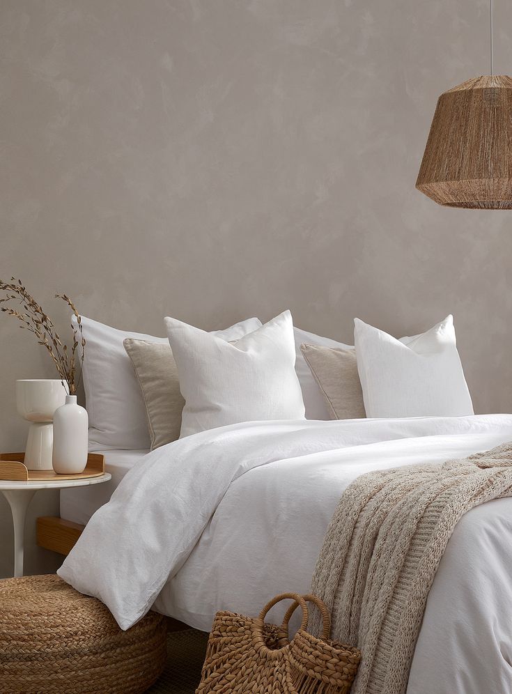 a bed with white linens and pillows in a neutral colored bedroom, next to a wicker basket