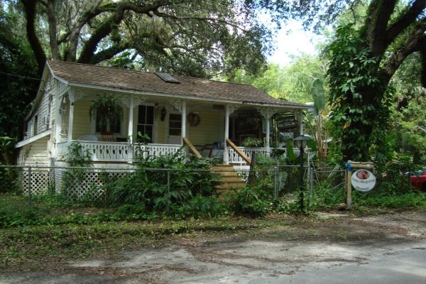 a small white house sitting next to a lush green tree filled forest in front of a red car